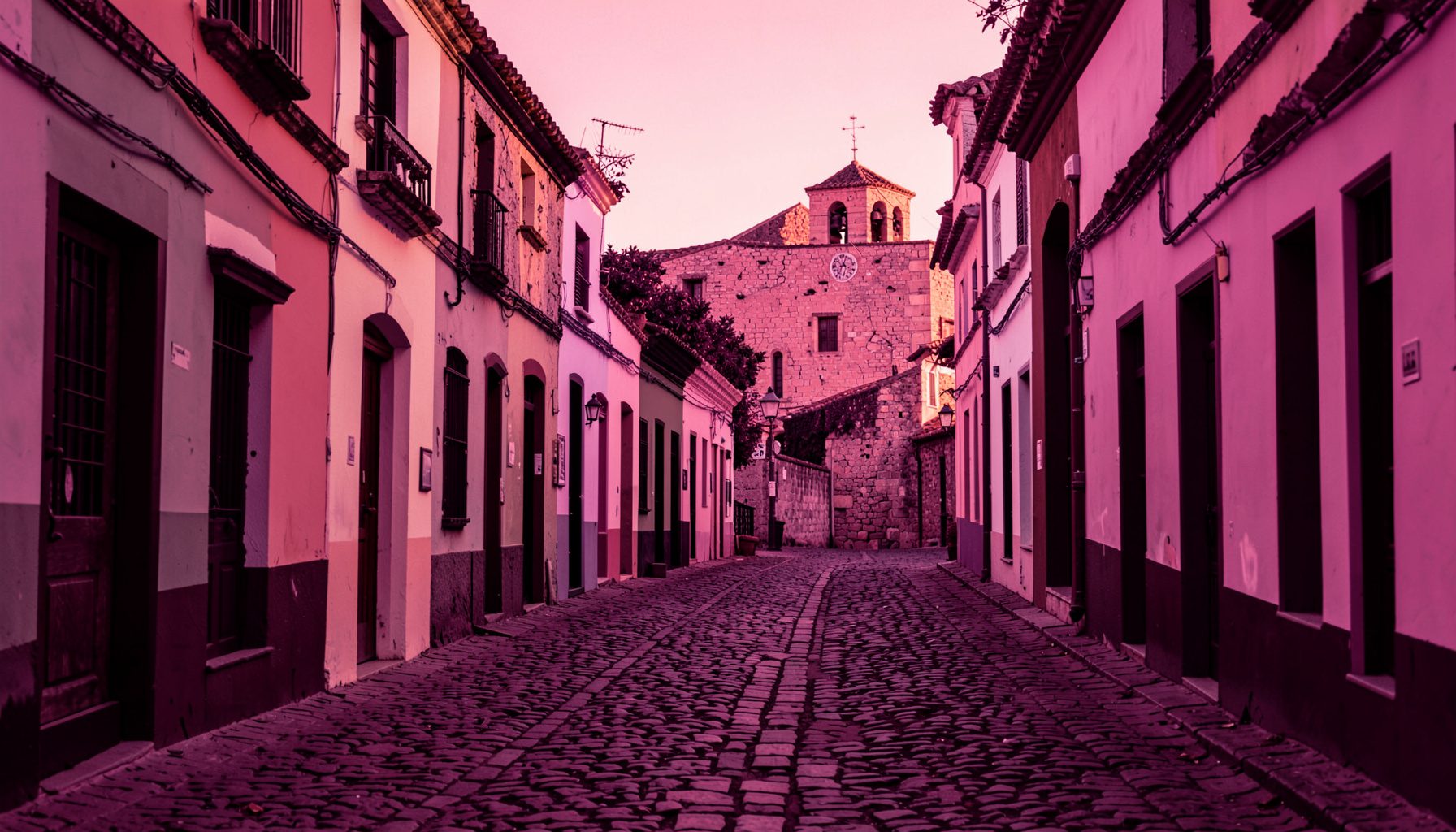 Calle empedrada con fachadas coloniales de colores cálidos y una iglesia de piedra rosada al fondo, bajo la luz dorada del atardecer.