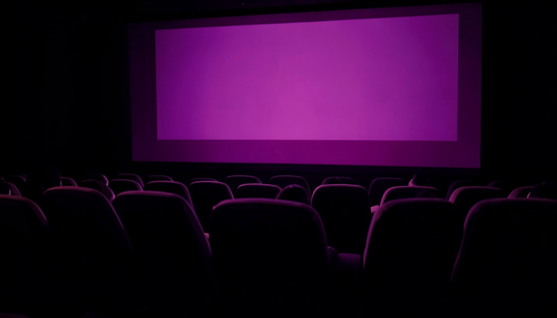 Silueta de butacas de cine iluminadas por una pantalla en penumbra, duotono violeta y dorado.